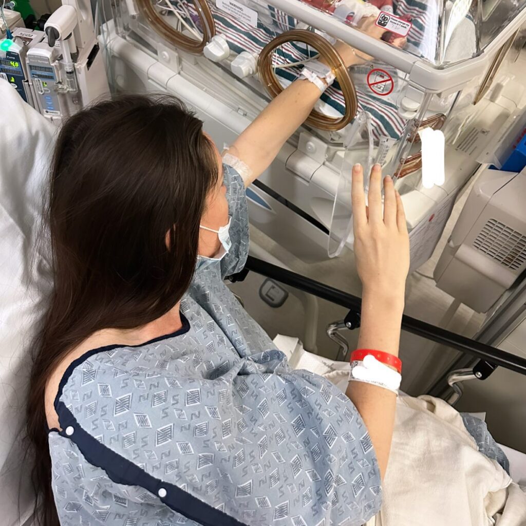 Image of a woman in the hospital touching the baby incubator next to her. 