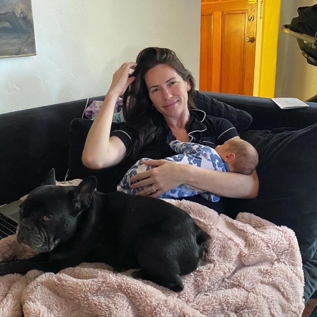 Image of woman on a couch holding her baby and having her dog besides her. 