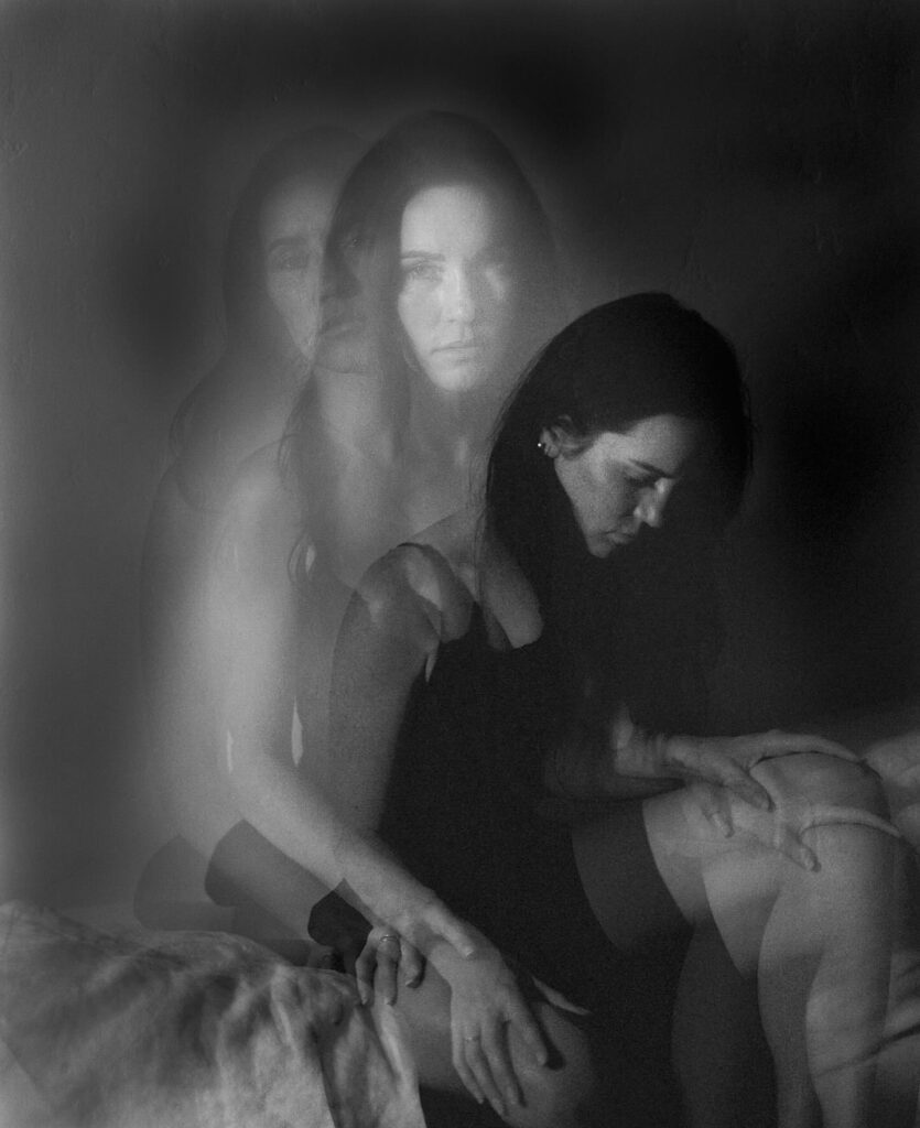Black and white image of woman in a dark room sitting on bed. 